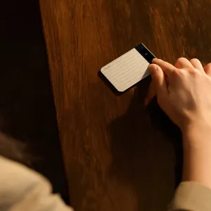 Grabadora de voz PLAUD Note Pro sobre una mesa de madera, con una mano tocándolo, mostrando el cuerpo plateado texturizado y el panel de pantalla negro
