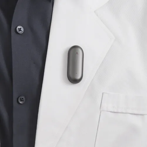 Längliches graues Gerät mit abgerundeten Kanten und einer kleinen Anzeige, am Revers eines weißen Blazers über einem dunklen Hemd im Brustbereich befestigt.